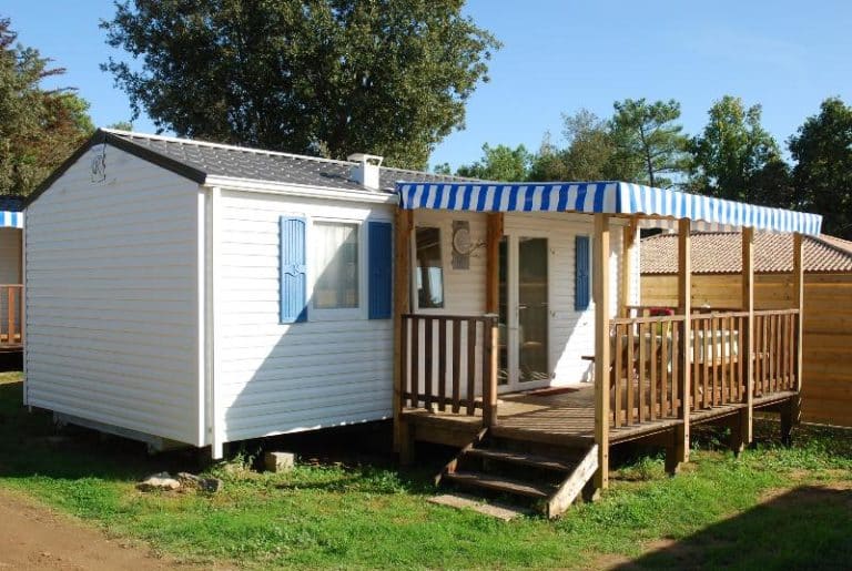 Mobil-home blanc avec terrasse en bois, entouré d'arbres.